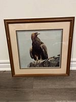 Front view of framed golden eagle print showing bird perched on rocks within cream matting and brown frame on a light colored wall.