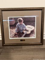 Framed print showing the full frame, matting, image of a boy with guitar, placed on floor, visible wooden frame and plaque