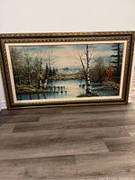 Front view of large oil painting on canvas depicting a cabin in woods by pond with mountains in background, framed in an ornate vintage wooden frame.