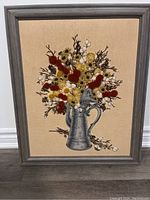 Front view of framed needlepoint embroidery featuring a bouquet of wild flowers in a gray pitcher.