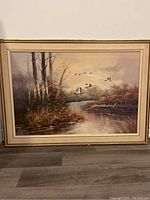 Full view of the vintage oil painting depicting ducks flying over a river with surrounding trees and grasses, framed.