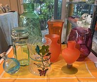 View of all 9 glass items arranged on a yellow cloth: includes green vase, amber and blue decanters with stoppers, three orange glass pieces, purple pitcher, small glass jar with wire lid, and clear decorated tumbler.