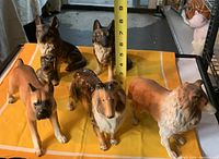 Top view of six dog figurines on yellow cloth with measuring tape showing height up to 9 inches, includes Boxer, Collies, and German Shepherds