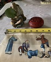 The Mudman ceramic figurine seated holding a leaf fan next to the round red cinnabar trinket box, both shown with a measuring tape indicating size approximately 6 inches long.