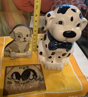 Front and top view of Dalmatian cookie jar, standing puppy plaque, and rectangular Welcome dog plaque on a yellow cloth with measurement markings.