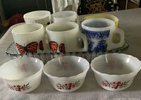 Photo showing six Fire King mugs (two butterfly print, one Asian Blue Willow, one yellow, one C-B'er titled, and one milk glass) arranged in and around the clear glass baking dish, along with three small floral dessert cups in front.