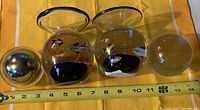 Four glass art pieces arranged in a row with measuring tape showing sizes approx. 4 to 5.5 inches wide. Two fish aquarium paperweights with clear rounded tops and two glass spheres with internal elements.