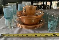 Photo showing peach luster Fire King Ware including mug, plates, bowls, dessert cups with retro aqua palm tree and aqua swirl juice glasses with tape measure for scale.