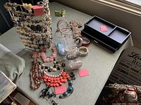 Photo of layout including hanging plexiglass or metal necklace stand, multiple necklaces with beaded and stone designs, bracelets, and black jewelry box on table