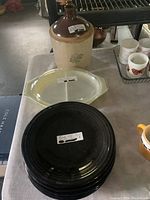 Photo showing 1 gallon Western Stoneware Co jug, yellow Pyrex divided dish with clear lid, and stack of black Fiesta plates with one cobalt blue plate beneath.