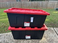 Two stacked black Hyper Tough storage totes with red lids placed outdoors on concrete and grass.