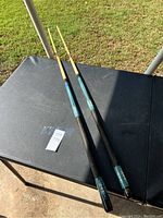 Two pool cues laid on a black table outdoors on a sunny day showing turquoise grip sections and black textured handles, with wooden tips.