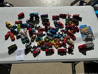 Overhead view of numerous die-cast toy cars in various colors and models scattered on a table, including one Hot Wheels carded car.