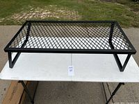 Black metal camping grill on white folding table, showing the grill with legs extended for use, rectangular shape with expanded mesh surface.
