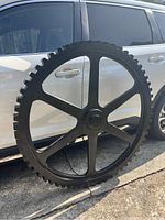 Large black wooden gear wheel leaning against white vehicle outdoors under sunlight, showing full detail of gear teeth and spokes.
