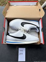 Photo of white Nike Cortez shoes with black swooshes inside open shoebox showing side profile and interior details
