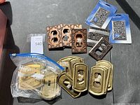 Overview of various decorative metal faceplates of brass, bronze, and dark finishes along with some packaged sets.