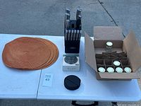 Overview photo showing brown woven round coasters stacked, knife set in black block with 7 knives, small black object (possibly a container), and partially visible box of jars.