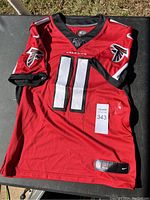 Front view of red Atlanta Falcons football jersey with number 11 and Falcons logos on sleeves, displayed on table.