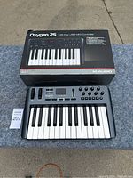 M-Audio Oxygen 25 MIDI Controller with its original box on a gray surface outdoors.