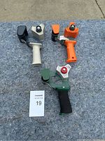 Three handheld tape dispensers photographed on concrete surface, showing colors black/beige, orange, and green with black handles.