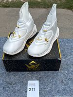 Front and side view of white Tronus high-top athletic shoes on top of original box outdoors.