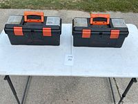Two closed black plastic tool boxes with transparent lid compartments, orange latches and handles on a white folding table outside.