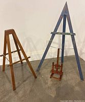 Three easels side by side: tall blue sitting field easel (42 inches), medium natural wood display easel (31 inches), small reddish wood tabletop easel (18 inches).