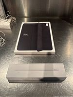 Boxed Apple iPad Smart Folio case and boxed Apple Pencil on a metal surface.