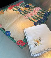 Floral appliqué placemat with bright colors of flowers and green stems on a light fabric background, alongside folded white embroidered napkins with floral design and scalloped edges.