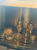 Photo showing full silver plate barware set with cocktail mixer, water pitcher, two metal cups, condiment jar, coasters, nut bowl, ashtray, and two crystal glasses on metal surface.