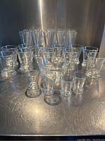 Set showing 11 tumblers (back row), 10 parfait glasses (middle), 1 bowl, 6 mini pudding cups/shot glasses (front) arranged on metal surface with uniform background.