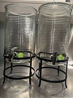 Two clear ribbed glass beverage dispensers side by side on black metal stands, one with a lid and one without.
