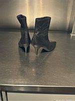 Two black leather ankle boots, viewed from rear angles showing heels and backs. Positioned on metal surface under warm lighting.