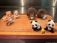 Top down photo of all seven Calico Critters animal figures on a wooden surface showing 2 hedgehogs, 3 kitties, and 2 pandas with scarves and honey pot accessory.