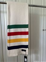 Blanket hanging showing off-white base color with green, red, yellow and navy stripes and a brand tag attached by a string.