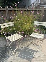 Two white/cream painted iron outdoor garden chairs with wooden slats on seat and backrest, placed on stone patio with flowering plants in the background.