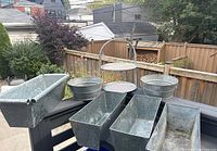 View showing 4 galvanized rectangular pots, 2 round galvanized buckets or tubs, and a large light aluminum rectangular planter on a surface outdoors, with a fenced backyard in the background.