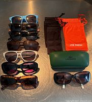 Eight pairs of sunglasses arranged in two columns, six black cloth bags plus one orange Joe Fresh cloth bag, and a green textured hard Lacoste sunglasses case with one pair of black/white Lacoste sunglasses.