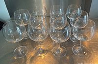 Eleven clear Riedel wine glasses placed on a metallic surface in three rows, showcasing their large, rounded bowl shape and long stems.
