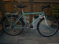 Side view of turquoise Rocky Mountain Fusion bicycle on tiled floor near wooden fence, showing rack, fenders, tires, seat, and handlebar grips