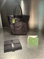 Black French Francis tote satchel with two handles and attached green tag, positioned behind a black wallet with leather strap detail on a metal surface