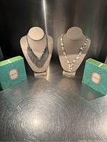 Two necklaces displayed on beige linen stands with two turquoise boxes on either side on a metal surface.