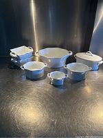 Front view of white ceramic serving dishes on a metal surface with reflections; seven pieces total including round and square scalloped dishes and a covered casserole dish.