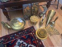 Full lot overview showing various brass items arranged on wooden floor and carpet, including dishes, figurines, plaques, horn and bell.