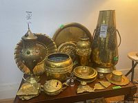 Photo showing assortment of brassware items on table including tall vase, urn, bowls, dishes, and lidded containers.