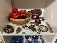 Overall shelf showing assortment of costume jewelry including red bangles in basket, assorted brooches, pendants, compact and cosmetic mirrors, and fabric pouch.