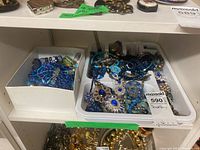 Two plastic trays containing various blue, green and gold tone necklaces and bracelets
