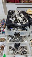 Costume jewelry organized in clear plastic drawers showing necklaces on top and various metallic bangles, bracelets, and rings inside drawers, along with a watch and colorful beads visible on side.