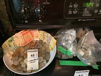 Bowl containing assorted international coins and several international banknotes including Mexican pesos, surrounding three plastic bags tagged 'US Coins' filled with assorted US coins.
